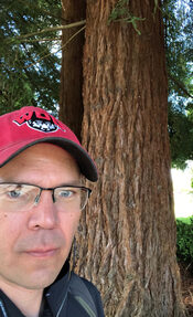 A man in a red cap and glasses stands before a redwood tree.