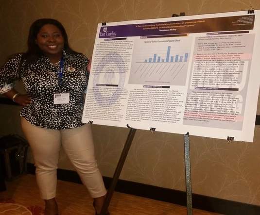 A Black woman in a printed shirt and light pants smiles at the camera. She stands by a conference poster.