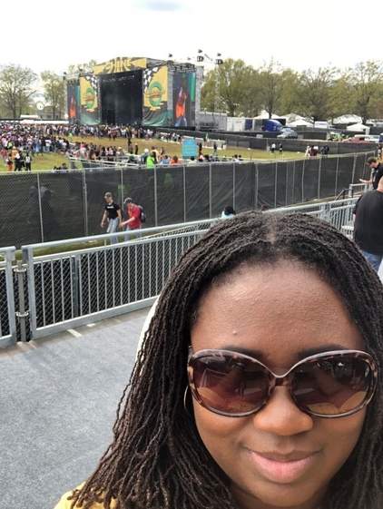 A Black woman in sisterlocs and wearing sunglasses smiles at the camera. Behind her stands a festival stage.