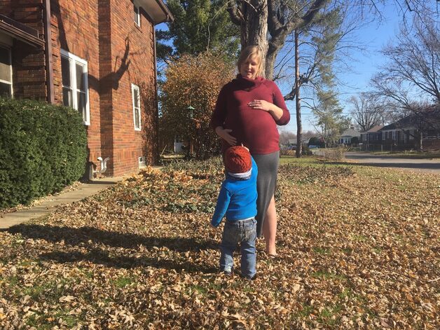 A white pregnant woman wearing a red turtleneck stands outside on a fall day with a small child in a blue sweater and red cap.