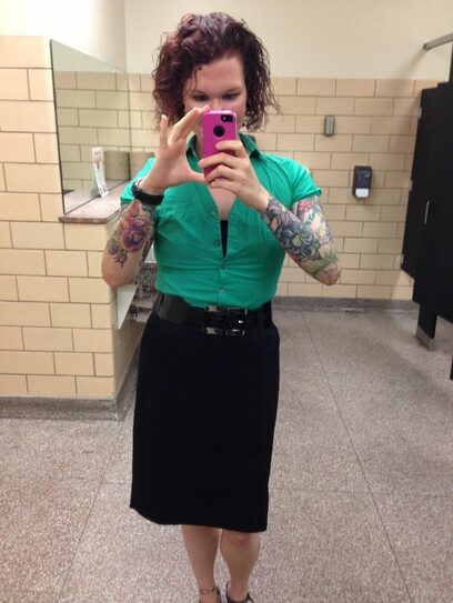 A redhaired white woman in a green blouse and black skirt takes a mirror selfie.
