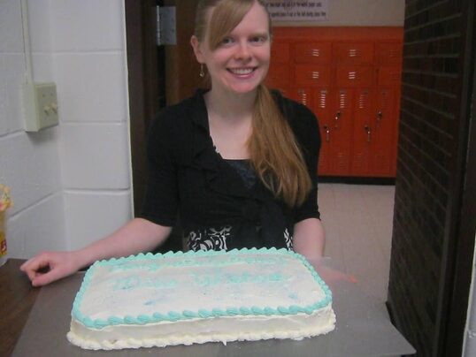 A blonde white woman in a black dress smiles at the camera. Before her on a table is a white iced cake with blue decoration.