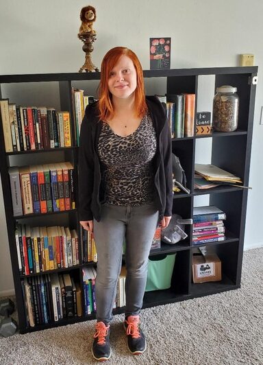 A redhaired white woman dressed in a print shirt, black sweater, and jeans smiles at the camera.