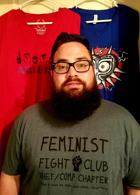 Man wearing a t-shirt reading “Feminist Fight Club Rhet/Comp Chapter.” Behind him are two other screen printed t-shirts.