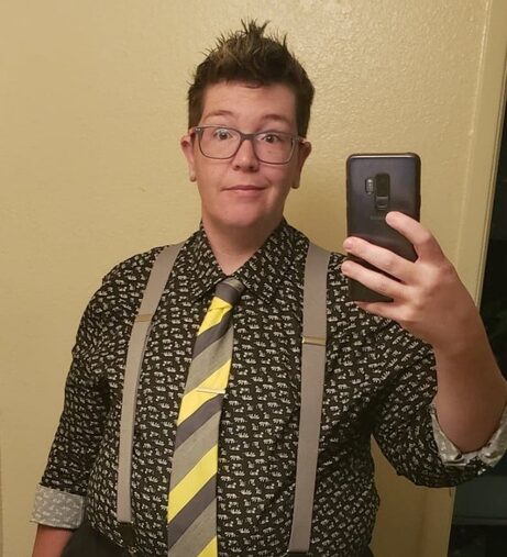 Griffin stands in front of a mirror holding up a phone to take a selfie. They are wearing a black patterned button-up dress shirt rolled up to the elbows, a yellow and grey striped tie, and grey suspenders. They are wearing a bemused and questioning facial expression.
