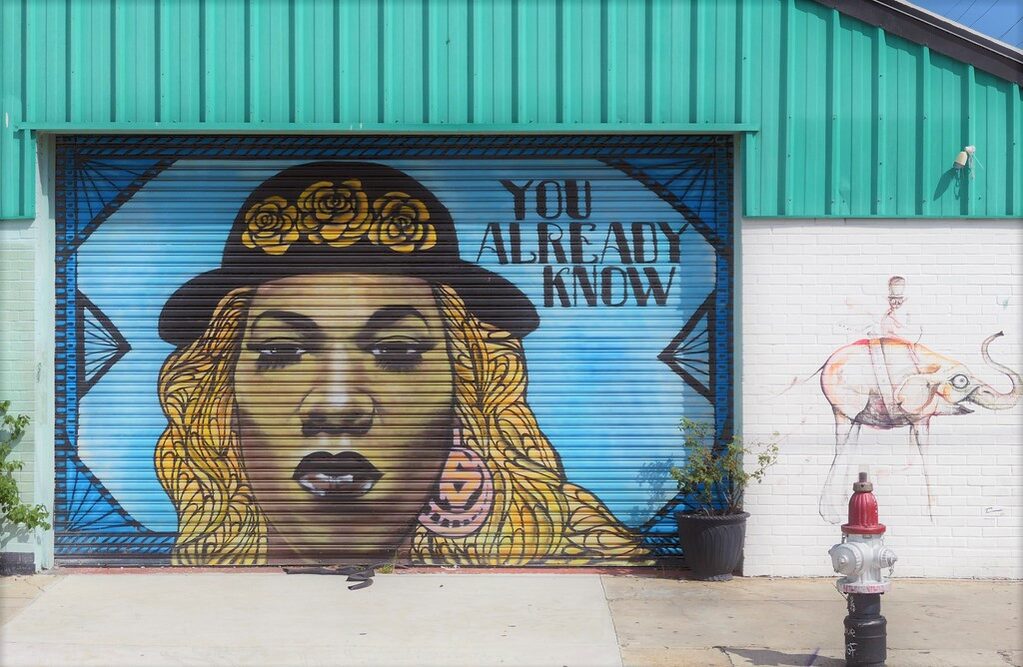 A street mural of Big Freedia wearing a black hat with yellow flowers. It reads 